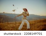 Fashionable woman wearing stylish autumn outfit with orange hat, sunglasses, white wide leg jeans, checkered blazer, belt, walking in mountain nature during the sunset. Copy, empty space for text