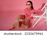 Fashionable confident woman wearing trendy 70s style outfit with big sunglasses, pink turtleneck top, sequined flare trousers, golden  shoes. Full-length studio portrait. Copy, empty space for text
