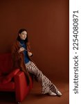 Small photo of Fashionable confident woman wearing trendy blue turtleneck, brown faux fur coat, chunky chain, flared trousers with zebra print, white cowboy boots. Studio fashion full-length portrait.