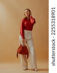 Small photo of Happy smiling fashionable woman wearing elegant satin blouse, white wide leg trousers, holding trendy suede fringed bag, posing on beige background. Full-length studio fashion portrait