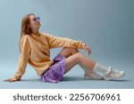 Fashionable confident girl wearing purple sunglasses, pleated mini skirt, yellow hoodie, socks, high top colorful sneakers, sitting, posing on blue background. Full-length studio portrait. Copy space