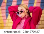 Fashionable confident blonde woman wearing trendy pink sunglasses, fuchsia color coat, turtleneck, blowing bubble with chewing gum, posing outdoor