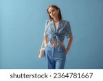 Fashionable young woman wearing  knotted chiffon blouse, trousers, carrying trendy white leather padded cassette shoulder bag, posing on blue background. Studio portrait. Copy, empty space for text