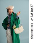 Small photo of Fashionable happy smiling woman wearing beret, elegant green coat, turtleneck, layered pearl necklace, holding trendy white leather purse with spring bouquet of snowdrops, posing on blue background