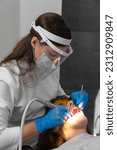 Small photo of Female dentist and assistant removing dental calculus from teeth. Visit is in proffessional dental clinic. Woman sits on dental chair. Drilling and treatment of tooth, filling. Vertical photo