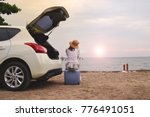 A young woman driving a car to beach . Back view of young girl sitting on blue travel bag and looking to the seascape against the sunset.