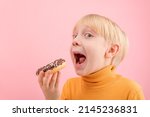 Small photo of Towheaded boy wants to bite donut with chocolate icing. Close up portrait of child with doughnut on pink background