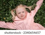 Small photo of Close up portrait of Caucasian blonde girl with two ponytails in pink jacket on green grass. Towheaded small girl outside