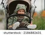 Small photo of A female toddler is eating the safety bumper bar of her stroller on a cloudy day. A young girl in a baby carriage in a village green.