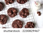 A tray of gooey triple chocolate cookies sit on a silver cooling tray next to a glass bottle of milk.  A striped straw emerges from the milk bottle, with two chunks of chocolate next to it.