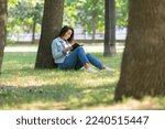 Small photo of Digital detox concept - Asian woman reading a book in the park. She sitting on the grass under the tree, reads a book and takes a break from constantly working at the computer and constantly being