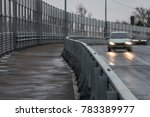 A wide pedestrian road is left along the high-speed viaduct. Silhouettes of two cars on the road. Powerful anti-noise protective fences along the highway.