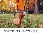 Orange knee high boots. Fashionable woman wearing stylish blazer plaid mini skirt walking in fall park with beige handbag among leaves. Copy space