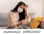 Small photo of Young Asian woman wearing face mask have a cold and high fever while checking body temperature by using digital thermometer. Daily lifestyle health care concept.
