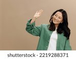 Happy cheerful Asian student young woman in khaki green shirt greet someone waving hand posing isolated on over beige pastel studio background. Cool fashion offer. Lifestyle and Emotions concept