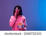 Small photo of Shocked awesome brunet woman in pink hoodie trendy specular sunglasses eat popcorn open mouth posing isolated in blue violet color light background. Neon party Cinema concept. Copy space