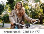 Smiling woman with curly hair in a coat rides a bicycle in a sunny park. Outdoor portrait. Beautiful woman enjoys nature. Lifestyle. Relax, nature concept. 