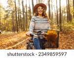 Smiling young woman in a hat and a stylish sweater with a bicycle walks and enjoys the autumn weather in the forest, among the yellow leaves at sunset.