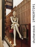 Small photo of white plastic mannikin figure of woman in black top sits in showcase shop window under bus stop sign in city, concept of soulless people, public life. Selective focus, film noise