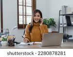 Smiling woman sitting in an home writing down notes talking from her telephone and laptop on the table