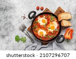 Small photo of Shakshuka in a frying pan on a gray rustic background. Jewish scrambled eggs. Poached eggs in a spicy tomato pepper sauce. Top view, flat lay. Textured object, selective focus.