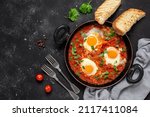 Small photo of Shakshuka eggs in a pan with toast on a black concrete background. Poached eggs in a spicy tomato pepper sauce. Traditional Jewish scrambled eggs. Top view, flat lay. Textured object, selective focus.