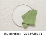 Green linen napkin on a clean beige plate, beige textile background. Top view, flat lay.