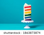 Single slice of birthday cake on a white cakestand minimalist on a blue background. Homemade cake with multicolored layers and buttercream