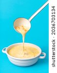 Small photo of Delicious greek soup in white enamel bowl isolated on a blue background. Pouring soup from an enamel ladle into the bowl.