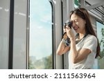 A Beautiful Asian female tourist with camera in the passenger cabin, traveling by sky train, taking snapshot photos when transporting in urban view, city lifestyle by railway, happy journey vacation.