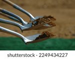 Close up of five shovels filled with dirt during a ground breaking ceremony. A few shovels are in focus and the rest are blurred against a brown and green background.