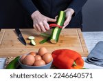 Small photo of Working woman's hands peeling courgette on wooden kitchen board. Faded in front, grey bowl with eggs, red pepper and another courgette. Concept of food preparation. Selective focus