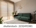 Elegant living room with big window wall behind wooden blinds, comfortable green sofa, carpet and decorative gold wall lamp