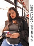 Small photo of Pregnant Latina woman traveling on an urban bus while looking at her cell phone, red colors predominate on the bus