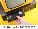 Small photo of VHS videocassette is put into the video recorder to watch the video. Old yellow vintage TV with VCR and videotape on black background from 1980s, 1990s, 2000s.