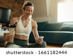 Happy sportswoman using smart phone while drinking protein shake after a workout at home. 