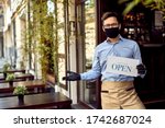 Happy waiter with protective face mask holding open sign while standing at cafe doorway. 