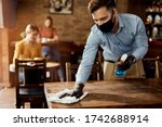 Small photo of Young waiter wearing protective face mask while cleaning tables while working in a cafe.