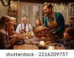 Small photo of Happy man bringing roast turkey at the table during Thanksgiving dinner with his multigeneration family.
