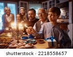 Small photo of Happy extended Jewish family celebrating Hanukkah while gathering at dining table. Focus is on boy lighting candles in menorah.