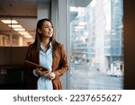 Young businesswoman holding digital tablet and day dreaming while looking through the window from her office. Copy space.