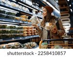 Smiling woman reading label on food package while buying groceries from refrigerated section in supermarket.