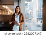 Young happy businesswoman using digital tablet while standing by the window in the office and looking at camera. Copy space. 