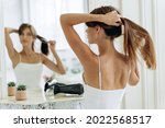 Young confident woman wearing white domestic clothes looking at her reflection while sitting at the mirror and brush long healthy brunette hair. Millennial female doing ponytail at the morning 