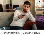 Small photo of Photo of young bored man sitting on couch at home and changing TV channels with disinterest. Stock photo