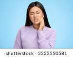 Small photo of Portrait of unhappy teen girl in hoodie touching sore cheek, frowning from acute pain, suffering cracked teeth, gum recession, dental problems. Indoor studio shot isolated on blue background
