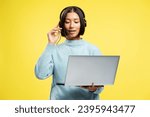 Attractive beautiful Asian businesswoman, saleswoman holding laptop, wearing headphones, talking, answering call standing isolated on yellow background. Contact center concept