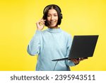 Portrait of cheerful smiling Asian woman, call center operator holding laptop, wearing headphones, looking at camera. Young female employee isolated on yellow background. Online communication