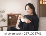 An Asian woman is sitting on the sofa, holding a cup and drinking soup. She is wearing long sleeves.