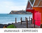 View of cliffs and Marine Drive, Scarborough from the colourful, orange and red beach huts in the North Bay. Roof tops of lower  huts are visible. Sea between the huts and cliffs.